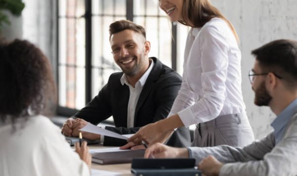 Happy diverse colleagues having fun at corporate meeting, sitting at table in modern boardroom, smiling friendly businesswoman leading briefing, business partners discussing sharing ideas